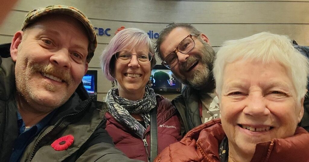 Selfie photo of two women and two men smiling, all wearing outdoor coats, in the background it appears to say CBC Radio, though the writing is partially obscured