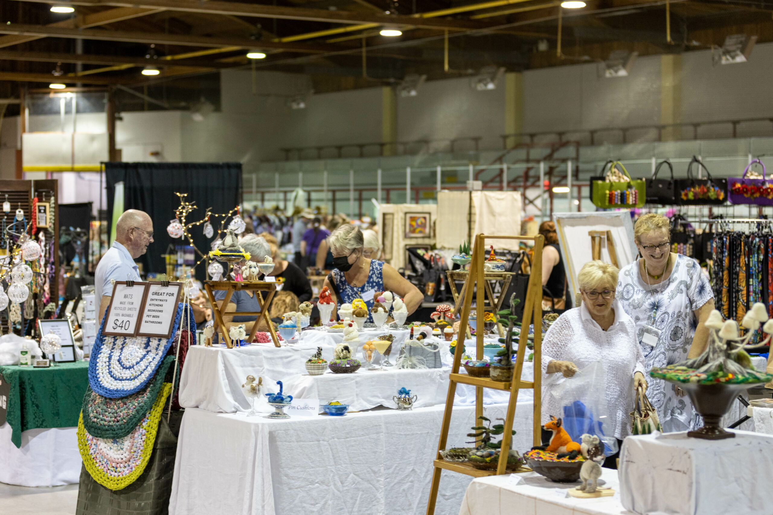 People in a large room at a market or event with many tables covered in white cloth, and many kinds of colourful object for sale including bags, ceramics, and fabric mats
