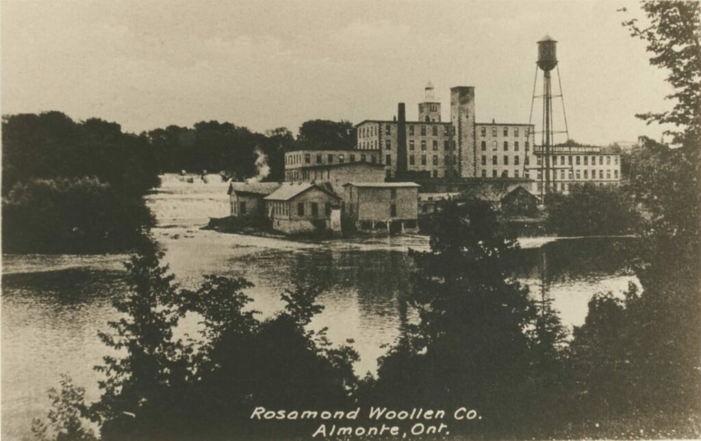 A sepia photo of a view across water to an old industrial factory complex, with a water tower, Rosamond Woollen Co. Almonte, Ont. hand-written at the bottom of the photo
