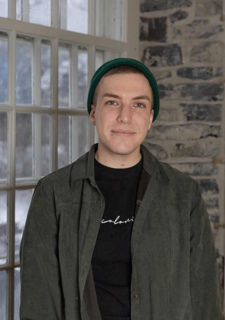 Colour photo of Maybe McInnis wearing a opened green button up on top of a black shirt, against a background of a grey stone wall and a window with small panes