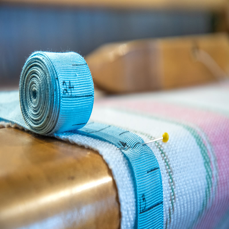 Close-up of a partially-unrolled blue measuring tape, pinned by a pin with a yellow top to white, green and pink-striped fabric